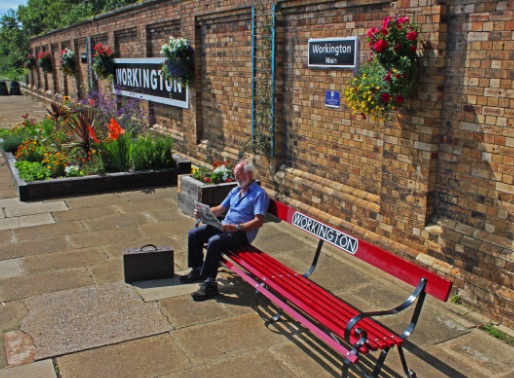 A photo of a man sitting at a platform bench, reading a newspaper, at Workington Train Station